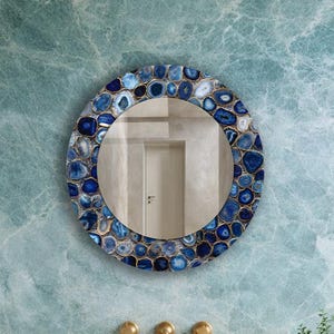 Puede incluir: Espejo redondo con borde de mosaico de piedras azules y blancas, colgado sobre un lavabo en un ba&ntilde;o. El mueble del lavabo es de madera clara, con encimera de piedra y lavabo sobre encimera. La pared es de color azul claro.