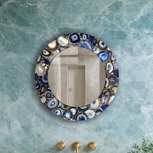May include: A round mirror with a decorative frame made of blue and white agate stones hangs above a bathroom vanity. The vanity has a light-colored countertop, a wooden cabinet, and a stone sink. The wall behind the mirror is a light blue.