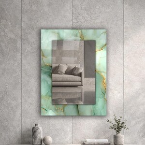 May include: A rectangular wall mirror featuring a decorative frame with a light green marble pattern and thin gold veining accents. The central reflective glass surface displays a clear reflection of a sofa and interior space.