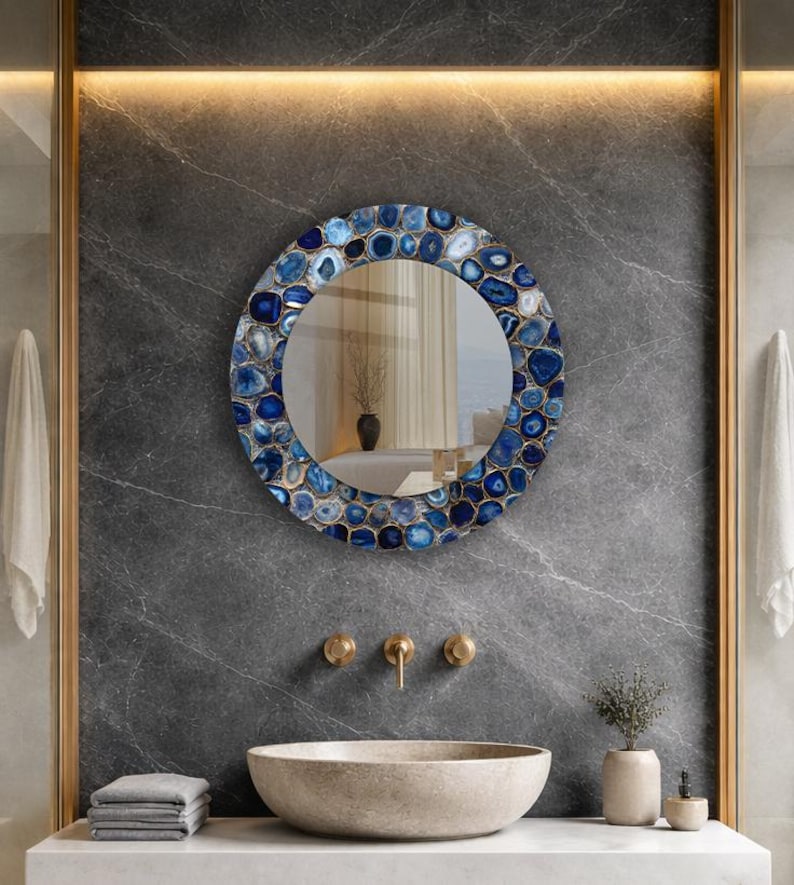 Puede incluir: Espejo redondo con borde de mosaico de piedras azules y blancas. El espejo est&aacute; montado en una pared de m&aacute;rmol gris sobre un lavabo de piedra con grifos dorados. Dise&ntilde;o de ba&ntilde;o moderno.