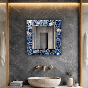 Puede incluir: Espejo cuadrado con marco de piedras azules y blancas, sobre una pared de m&aacute;rmol gris oscuro. El ba&ntilde;o cuenta con un lavabo de piedra, grifos dorados y una peque&ntilde;a planta en una maceta beige. El espejo refleja una puerta.