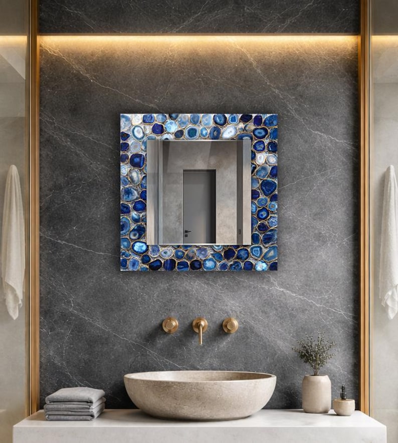 Puede incluir: Espejo cuadrado con marco de piedras azules y blancas, sobre una pared de m&aacute;rmol gris oscuro. El ba&ntilde;o cuenta con un lavabo de piedra, grifos dorados y una peque&ntilde;a planta en una maceta beige. El espejo refleja una puerta.