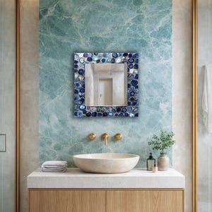 Puede incluir: Un espejo cuadrado con un marco de estilo mosaico con piedras azules y blancas. El espejo cuelga sobre una encimera blanca con un lavabo y grifos dorados. La pared detr&aacute;s del espejo es de color verde azulado veteado.