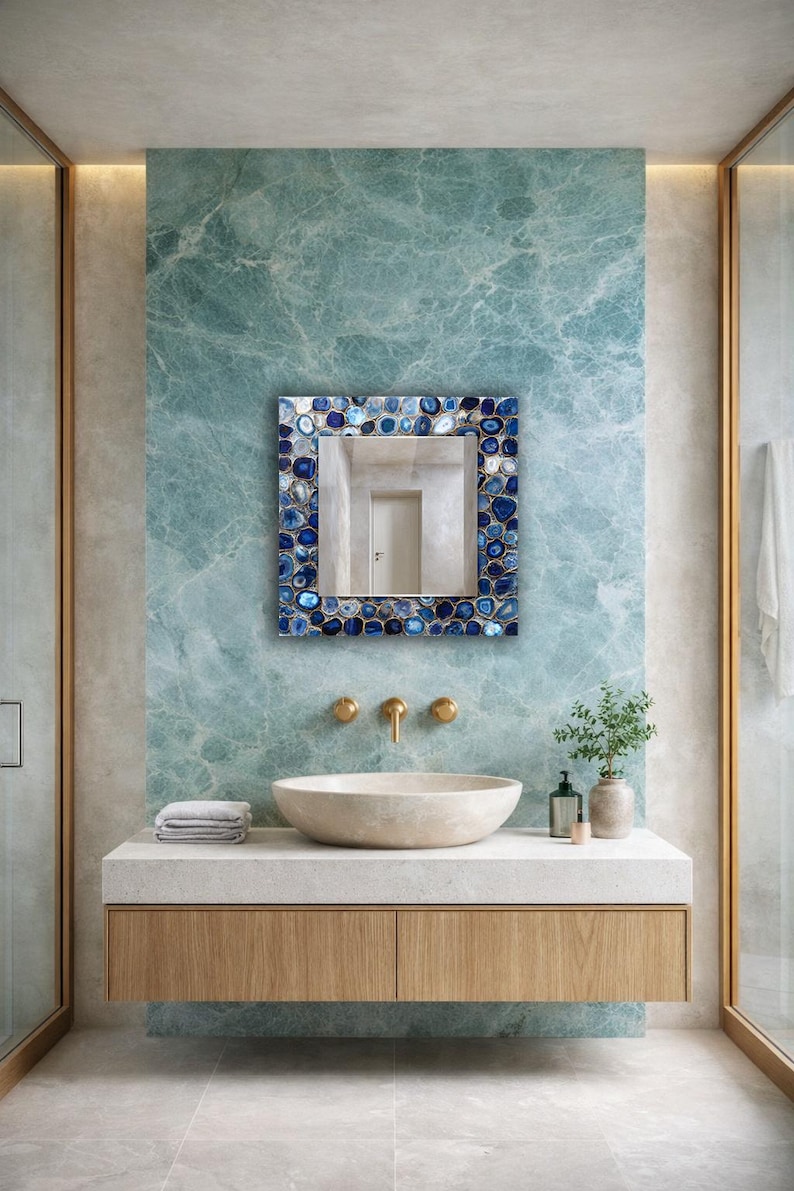 Puede incluir: Un espejo cuadrado con un marco de estilo mosaico con piedras azules y blancas. El espejo cuelga sobre una encimera blanca con un lavabo y grifos dorados. La pared detr&aacute;s del espejo es de color verde azulado veteado.