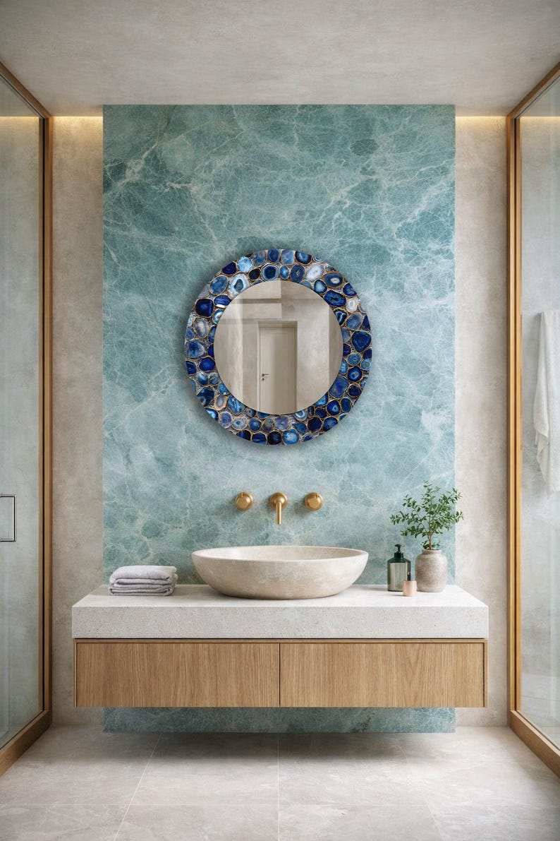 Puede incluir: Espejo redondo con borde de mosaico de piedras azules y blancas, colgado sobre un lavabo en un ba&ntilde;o. El mueble del lavabo es de madera clara, con encimera de piedra y lavabo sobre encimera. La pared es de color azul claro.