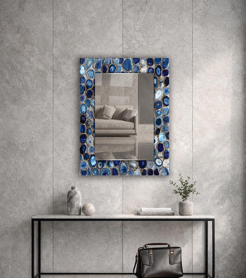 Puede incluir: Espejo rectangular con un marco adornado con piedras de &aacute;gata azules y blancas. El espejo refleja una habitaci&oacute;n con un sof&aacute;. El espejo est&aacute; montado en una pared gris sobre una mesa negra con objetos decorativos y una bolsa de cuero negra.