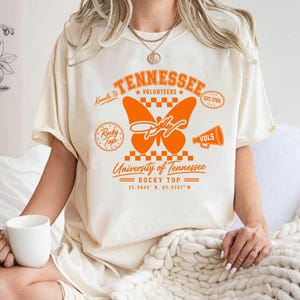 May include: A cream-coloured short-sleeve t-shirt featuring a large orange graphic on the front. The design includes a butterfly, a megaphone, a checkerboard pattern, and text reading Tennessee Volunteers, University of Tennessee, and Rocky Top.
