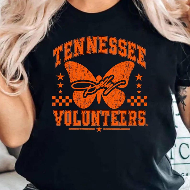 May include: A black t-shirt featuring a distressed orange graphic of a butterfly in the centre. The text "TENNESSEE" arches above the butterfly, while "VOLUNTEERS" is printed below. The design includes small stars and checkerboard patterns.