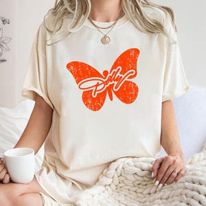 May include: A cream-coloured short-sleeve t-shirt featuring a large, distressed orange butterfly graphic on the chest. The butterfly design incorporates the word Dolly in a white cursive script across its wings. The shirt has a crew neckline.