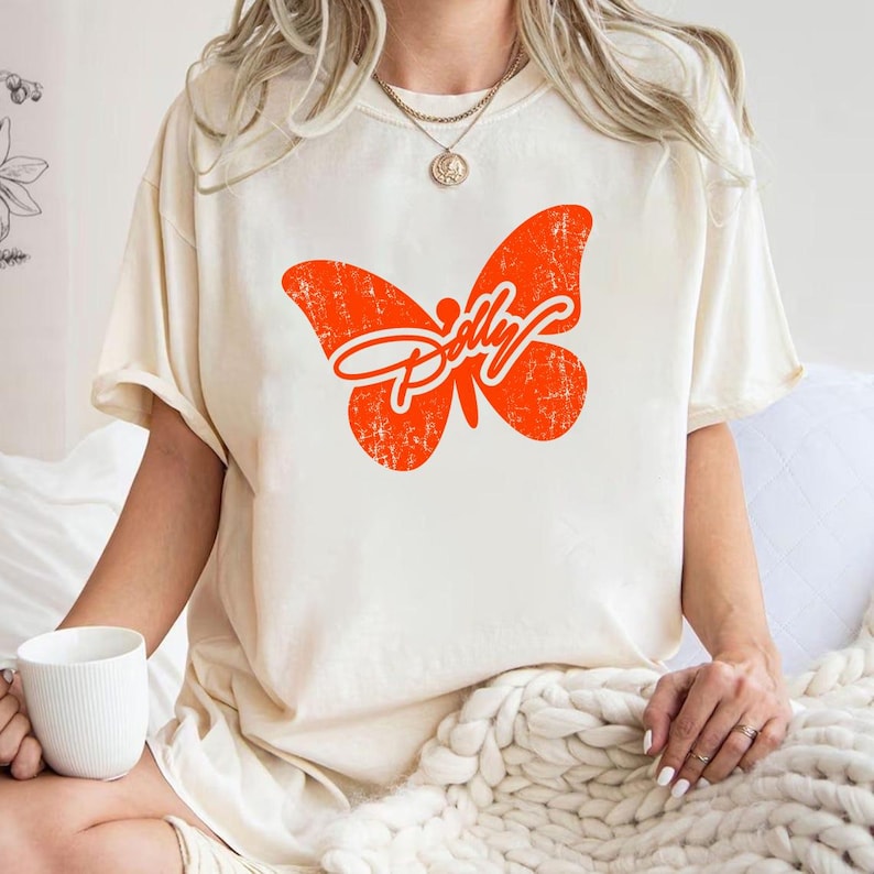 May include: A cream-coloured short-sleeve t-shirt featuring a large, distressed orange butterfly graphic on the chest. The butterfly design incorporates the word Dolly in a white cursive script across its wings. The shirt has a crew neckline.