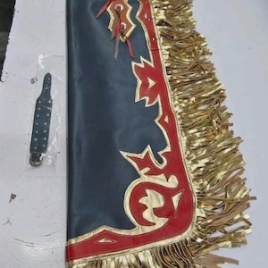 以下が含まれることがあります： 赤と金のアクセントと金のフリンジが付いた黒革のチャップス。チャップスは、縁に沿って装飾的なデザインが施されています。別の黒革ストラップが含まれています。