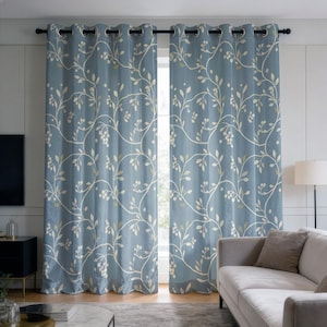 May include: A pair of floor-length curtains in a dusty blue shade featuring a repeating white vine and leaf pattern. The curtains are hung on a black rod with silver-toned metal grommets at the top, creating uniform vertical folds across the fabric.