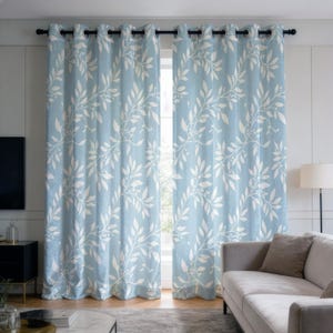 May include: A pair of light blue curtains with a white leaf and berry design. They are suspended from a black rod. The curtains frame a window. The room features a grey sofa and a black side table, with a neutral colour palette.