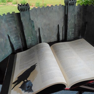 K&ouml;nnte beinhalten: Eine Schwarzwei&szlig;illustration eines Rabens, der auf einem Sch&auml;del sitzt, gedruckt auf der Seite eines gro&szlig;en, ge&ouml;ffneten Buches. Das Buch liegt auf einem Tisch vor einem grauen Pappschloss-Hintergrund.