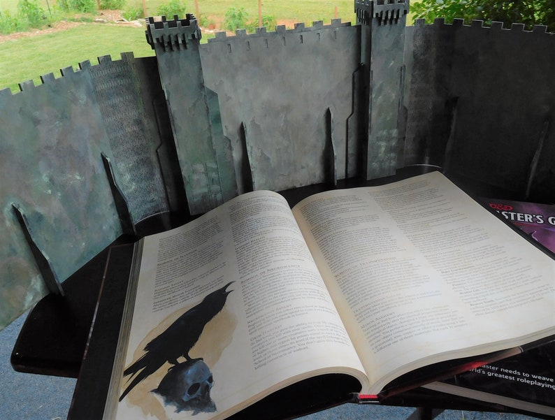 K&ouml;nnte beinhalten: Eine Schwarzwei&szlig;illustration eines Rabens, der auf einem Sch&auml;del sitzt, gedruckt auf der Seite eines gro&szlig;en, ge&ouml;ffneten Buches. Das Buch liegt auf einem Tisch vor einem grauen Pappschloss-Hintergrund.