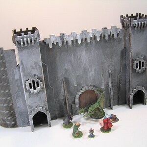 K&ouml;nnte beinhalten: Eine graue Steinburgmauer mit einer gro&szlig;en Holzt&uuml;r und einer gr&uuml;nen Drachenfigur davor. Vier Miniaturfiguren stehen vor der Burgmauer.