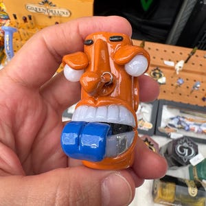 May include: A glossy orange glass figurine shaped like a stylized face with a prominent nose, white eyes, a silver nose ring, and a wide mouth showing white teeth and a protruding, thick, bright blue tongue that extends outward from the center.