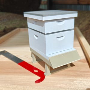 Puede incluir: Un modelo de colmena en miniatura de color blanco con techo gris y tabla de aterrizaje beige, sostenido por cuatro patas. Una herramienta de metal roja y plateada se sitúa junto a la colmena, sirviendo de referencia de escala.