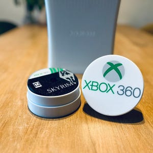 May include: An Xbox 360 console and two white circular coasters. One coaster features the Xbox logo and "XBOX 360" in green and gray. The other coaster has "SKYRIM" printed on it.