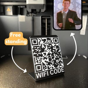 May include: A free-standing black and white WiFi code sign with a QR code. The sign is on a dark surface, with an image of a man in a black blazer and striped shirt in the background. The words "WIFI CODE" are printed on the sign.