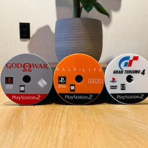 May include: Three PlayStation 2 game discs: "God of War" in gray and red, "Half-Life" in orange, and "Gran Turismo 4" in white and blue. Each disc has the PlayStation 2 logo and game title text. The discs are on a wooden surface.