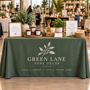 May include: A dark green tablecloth featuring the white logo and text GREEN LANE HOME DECOR, PLANTS, CANDLES, GIFTS, ARTISAN GOODS. On the table are eucalyptus, three brown jars, a reed diffuser, a candle, and signs reading NATURAL HANDCRAFTED HOME GOODS.