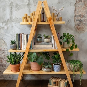 May include: Wooden ladder shelving unit with three shelves. The shelves display books, potted plants, candles, and decorative items. The unit is made of light-coloured wood and stands against a textured wall.