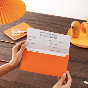 May include: An orange envelope holds a "SUPPORT WORKER INCIDENT REPORT" form. The scene includes an orange notepad, scissors, a yellow tray with a bird-shaped object, a cup with pencils, and a decorative orange lamp.