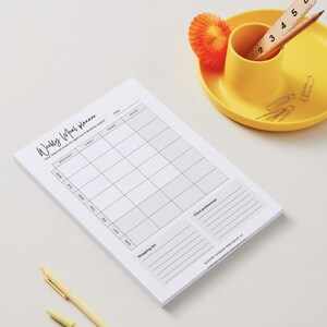 May include: A white weekly meal planner pad with a grid layout for daily meals. The pad includes sections for breakfast, lunch, dinner, and snacks. A yellow pen and a yellow dish with a pencil, ruler, and paper clips are also visible.