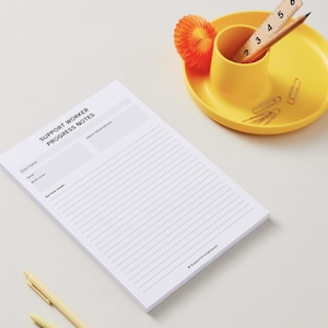 Pode incluir: Um bloco de notas branco com o texto "SUPPORT WORKER PROGRESS NOTES" está sobre uma superfície branca. Uma caneca e um prato amarelos contêm um lápis, uma régua, clipes de papel e uma decoração de papel laranja. Uma caneta amarela também é visível.