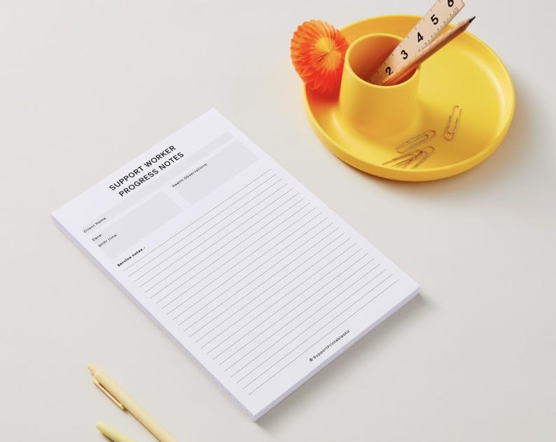 May include: A white notepad with the text "SUPPORT WORKER PROGRESS NOTES" is on a white surface. A yellow cup and plate hold a pencil, ruler, paper clips, and an orange paper decoration. A yellow pen is also visible.