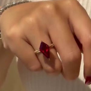 May include: A gold ring featuring a large, kite-shaped deep red gemstone set on a thin band adorned with small clear stones. The ring is worn on a finger, and a matching clear stone tennis bracelet is visible on the wrist.