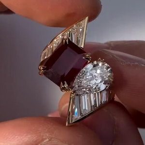 Puede incluir: Un anillo de compromiso con una piedra preciosa rectangular de color rojo intenso y una piedra clara en forma de pera, acentuado por piedras claras de corte baguette en un engaste de oro. El texto "No hook, just this engagement ring." es visible.