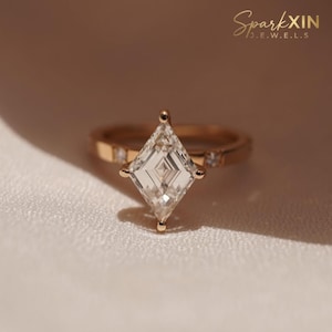 May include: A gold ring featuring a large, clear, kite-cut diamond. The band has small, embedded stones. The ring is set against a soft, white fabric. The text "SparkXIN JEWELS" is visible in the upper right corner.