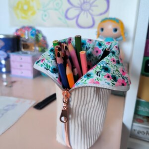 Puede incluir: Estuche de lápices de pana beige con forro floral, parcialmente abierto para revelar una colección de lápices de colores. La cremallera es de color oro rosa. Un dibujo de flores está en el fondo.