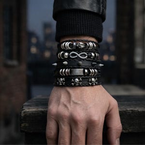 Può includere: Una pila di braccialetti in pelle nera con dettagli argentati e motivi a teschio, infinito e borchie. I braccialetti sono indossati su un polso, mostrando uno stile audace e audace. I braccialetti sono un mix di trame e design.
