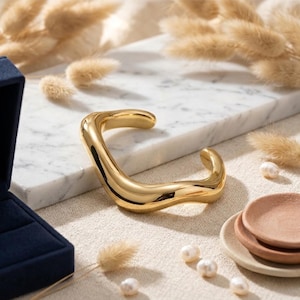Può includere: Un bracciale a polsino color oro con un design curvo e organico. Il bracciale è esposto su una superficie di marmo, con un portagioie blu e elementi decorativi sullo sfondo. Sono visibili anche perle e piattini.