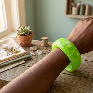 Può includere: Un braccialetto largo e traslucido verde lime con effetto marmorizzato è indossato sul braccio di una persona. Il braccialetto è il punto focale, posto su un tavolo di legno rustico. Altri oggetti sul tavolo includono una piccola pianta succulenta in vaso, cristalli e una bobina di filo.