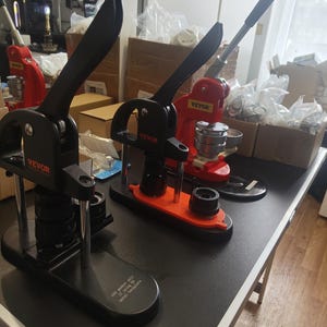 Peut inclure: Trois machines à fabriquer des boutons avec des composants noirs et rouges sont exposées sur une table noire. Les machines à boutons ont de longues poignées noires et portent la marque "VEVOR". Les machines à boutons sont dans un atelier.