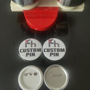 May include: A button maker with two custom pins. The pins are white with the text "CUSTOM PIN" in black. The button maker has a red handle and silver components. The back of the pins are silver and white.