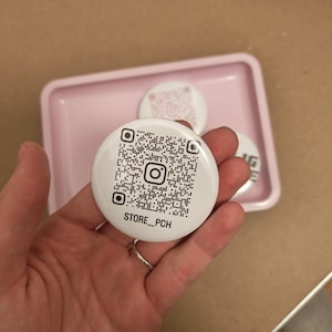 Könnte beinhalten: Weißer Button mit einem QR-Code und dem Instagram-Logo, mit dem Text "STORE_PCH". Zwei weitere Buttons befinden sich im Hintergrund, einer mit einem rosa QR-Code. Die Buttons liegen auf einem rosa Tablett.