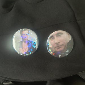 Puede incluir: Dos botones redondos y holográficos con imágenes de personas sobre tela negra. Un botón presenta a un hombre con traje, el otro a un hombre con camisa azul. Los botones tienen un efecto brillante e iridiscente.