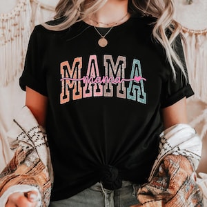 May include: Black t-shirt with the word "MAMA" in a colorful, ombre design. The word "mama" is written in a cursive font below the word "MAMA", with a small pink heart. The shirt is tied at the waist.