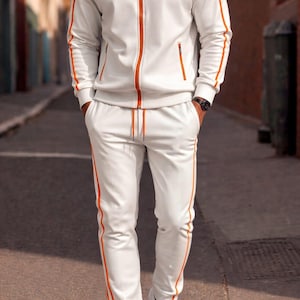 May include: White tracksuit with orange detailing. The jacket features a zip and pockets. The trousers have an orange stripe down the side. The outfit is completed with white trainers.