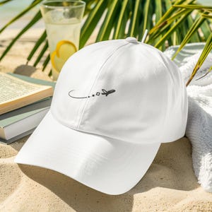 May include: White baseball cap with a curved brim, featuring a black airplane graphic and dotted flight path. The cap is set on a sandy beach with a drink and palm fronds in the background. A perfect accessory for travel.