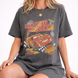Puede incluir: Camiseta gris oscuro con un dise&ntilde;o de Rayo McQueen de la pel&iacute;cula Cars. El dise&ntilde;o incluye el texto "Lightning McQueen" y "95", una bandera a cuadros y un trofeo. La camiseta tiene mangas cortas.