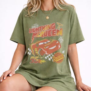 Puede incluir: Camiseta verde oliva con un dise&ntilde;o de Rayo McQueen de la pel&iacute;cula Cars. El dise&ntilde;o incluye el texto "Lightning McQueen" en rojo y amarillo, el n&uacute;mero "95" y la palabra "Rusteze". La camiseta tiene mangas cortas y un corte holgado.