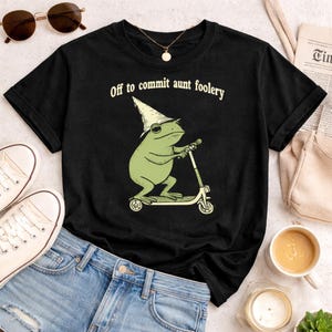 May include: Black t-shirt featuring a green frog riding a scooter and wearing a pointed hat. The text "Off to commit aunt foolery" is printed above the frog. The shirt is paired with blue denim shorts and white sneakers.