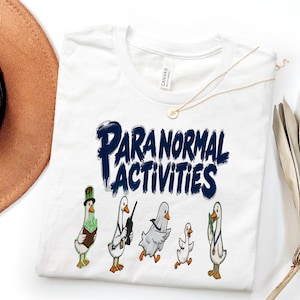 Puede incluir: Una camiseta blanca con el texto azul oscuro "PARANORMAL ACTIVITIES" sobre un gráfico de cinco patos de dibujos animados. Los patos aparecen en varios papeles, incluido uno con sombrero de copa y otro con una sábana de fantasma.
