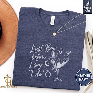 May include: A heather navy blue t-shirt featuring a white graphic of a ghost sitting in a martini glass, holding a shaker and a drink. The text reads "Last Boo before I say 'I do'" with a crescent moon and a diamond ring icon below the text.